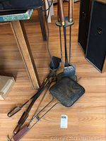 Photo showing fireplace tool stand with shovel, poker, brush and loose shovel, tongs, poker, and basket on wooden floor near table legs.