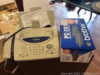 Full view of Brother IntelliFAX 1270e fax machine, setup guide, power cord, and two boxed Brother PC-201 printing cartridges on table