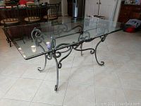 Glass top dining room table with ornate black metal base, viewed from a front angle to show top and metal scrollwork underneath.