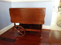 Side view of wooden dropleaf cart showing large wooden wheel (broken) and smaller caster wheel on floor.