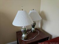 Pair of two lamps sitting on wooden table with cords visible. Bases are ceramic with painted medallions and gold accents. Shades are white and textured. Differing finials: clear crystal and green sphere.