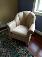 Upholstered armchair in cream fabric with rounded back and arms, visible staining on seat and arms, sitting on dark wooden floor with a patterned carpet nearby.