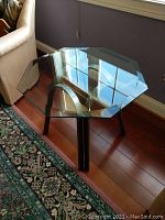 Slightly angled front-side view of the table showing glass top and metal legs with visible reflections and floor surface.