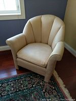 Front view of beige upholstered armchair with scallop-shaped padded back and cushioned seat on hardwood floor and patterned rug.
