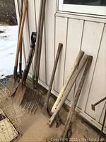 Photo showing collection of garden tools including shovels, pitchfork, and axes leaning against a wall on a concrete floor with snow outside.