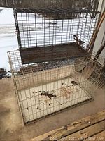Photo showing two animal cages, one white and one dark metal, placed outdoors on ground by a building.