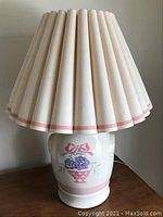 View of one porcelain table lamp with floral basket design on the base and pleated fabric shade with pink trim.