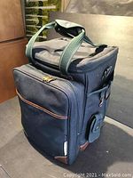 Closed navy blue cooler and picnic bag with green and white striped handles, accented with brown faux leather at base corners and side pockets. Label tag visible with brand 'Picnic by Ascot'.