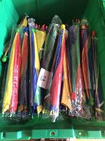 Twelve rainbow umbrella hats folded and stacked vertically in a green plastic bin, showing colorful panels and elastic bands visible in packaging.