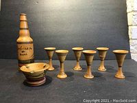 Hand-painted wooden bottle inscribed with text, six matching shot glasses and an ashtray arranged in a single line on dark surface with neutral background