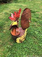Folk art metal chicken sculpture shown outdoors on grass, displaying front and side views to highlight painted details and rust on metal surfaces.