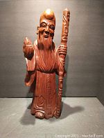 Front view of the tall carved wooden figure showing detailed facial features, flowing robes, holding a staff in one hand and a peach shaped object in the other.