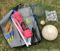 Complete set laid out on grass showing badminton racquets, net, stakes, shuttlecock, and balls.