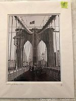 Black and white photograph showing Brooklyn Bridge, pedestrian walkway with people, suspension cables, NYC skyline. Photo has a border/mat. Signed or numbered in lower margin.
