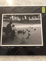 Framed and matted black and white photograph titled 'Feeding the Pigeons' showing the artist's signature and numbering 40/91 on the mat below the image. Shows the image of a person and child feeding pigeons on the sidewalk.