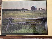 Color photograph depicting a rustic barn and weathered rail fence in a farm field. The scene includes green grass, some yellow wildflowers, and trees backing the barn under a cloudy sky.