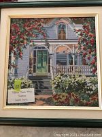 Framed cross stitch artwork showing a blue house with a porch and abundant flowers in the foreground, framed in a blue wooden frame with cream matting.