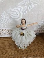 Front view of one Erika Dresden Art porcelain dancer figurine in white fabric skirt with lace trim on wooden surface in front of floral cloth background.