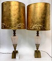 Pair of vintage table lamps with large gold-colored shades showing floral detail. Lamps have pink glass bodies with metal accents and square metal bases. Lamps connected with cords visible on floor.