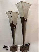 Pair of two metal base lamps with square, flared shades. One taller lamp with light-trimmed shade, one shorter lamp with dark-trimmed shade, cords attached.