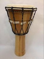 Front view of wooden djembe drum showing natural wood finish, off-white drumhead, and black rope tuning with plastic clips.