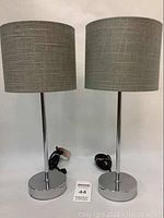 Pair of bedside table lamps showing chrome bases, chrome stems, and gray drum fabric lampshades, both plugged in with cords visible.