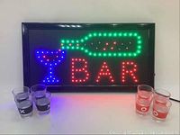 LED bar sign illuminated showing a green bottle, blue cocktail glass, and red 'BAR' letters with four shot glasses in front.