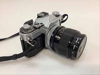 Three-quarter view of Canon AE-1 body with FD 35mm f/2 lens and neck strap attached, showing top dials and leatherette grip.