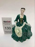 Front view of the green-dressed porcelain lady seated, holding beige hat with floral detail.