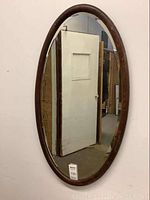 Front full view of large oval oak framed mirror hanging on wall reflecting adjacent door and room.