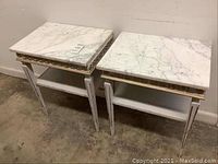 Two white marble top distressed end tables shown from a front angle on concrete floor near wall. Decorative gold trimming under marble surface, tapered legs, and lower shelf visible.