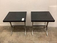 Front view of two square tables with black tops and brushed metal bases, showing their overall shape and size.