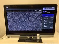 Front view of Sharp Aquos TV powered on showing static screen with on-screen input menu, stand attached, remote placed on stand