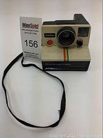 Front angled view of the Polaroid OneStep Land Camera showing lens, red shutter button, rainbow stripe, and black strap.