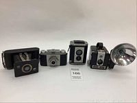 Four vintage cameras shown lined in a row: box camera with Twindar lens, Kodak Pony 135 model B, Brownie Reflex, Brownie Hawkeye with flash bulb reflector.
