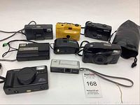 Seven assorted used point and shoot cameras displayed on a white surface including Minolta, Hanimex, Vivitar models and carrying pouch.