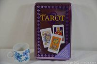 Rectangular purple tin with tarot card illustrations on lid next to a small coffee cup for scale