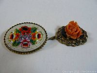 Full view of two brooches connected by a small chain. Oval micro mosaic floral brooch and round coral flower brooch with gold-tone frames.