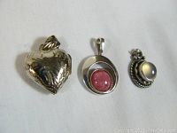 Three silver pendants against a white background showing front view of heart-shaped locket, circular pink stone pendant, and round moonstone-like pendant.