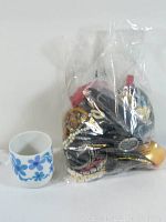 Photo of the lot showing costume jewelry inside a clear plastic bag next to a small white cup with blue flower pattern for size reference.