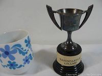 Vintage trophy with two handles and black wooden base with plaque reading 'Sandcastle Champ' next to a white mug with blue floral pattern for size comparison.