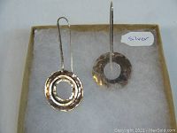 Top-down view of the two silver earrings in a box with a label 'silver'. One earring is concentric circles, the other is a single hollow circle.