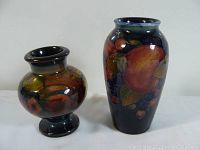 Front view of two Moorcroft vases, one taller and one shorter with bulbous body, both with fruit and leaf designs on dark backgrounds.