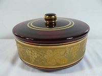 Front view of round antique lidded glass dish with gold grapevine decoration on maroon background and gold handle.