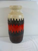 Front view of a tall pottery vase with beige upper section, textured black jagged mid section, and red and brown bands below.