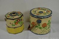 Two vintage ceramic cookie jars side by side. The larger one has a woven handle and orange floral decoration, the smaller one is shorter with a yellow bottom and floral design on the lid.