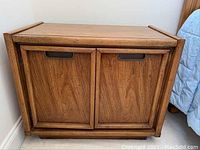 Two wooden nightstands constructed from wood with wood grain finish, showing general view of one unit with inset wood panel doors and recessed darker handles.