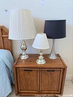 Photo showing three table lamps of different styles and sizes on a wooden nightstand next to a bed.