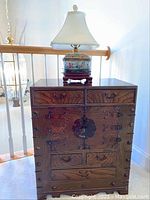 Front view of Asian style wooden cabinet with two top drawers, central cabinet doors with metal latch, and three lower drawers. Asian style lamp with fabric shade on top of cabinet.