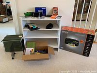 Photo showing white three-shelf bookcase, Epson Stylus NX420 printer box, office supplies including calculators, file separators, Pendaflex folders, sunglasses and black cases, and cardboard box with small office items on carpeted floor.